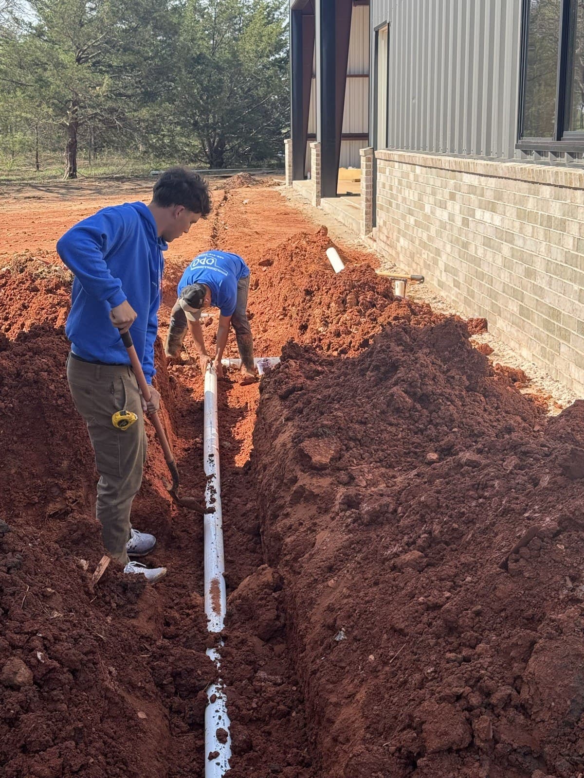 Two workers installing an underground plumbing line in a trench beside a building.